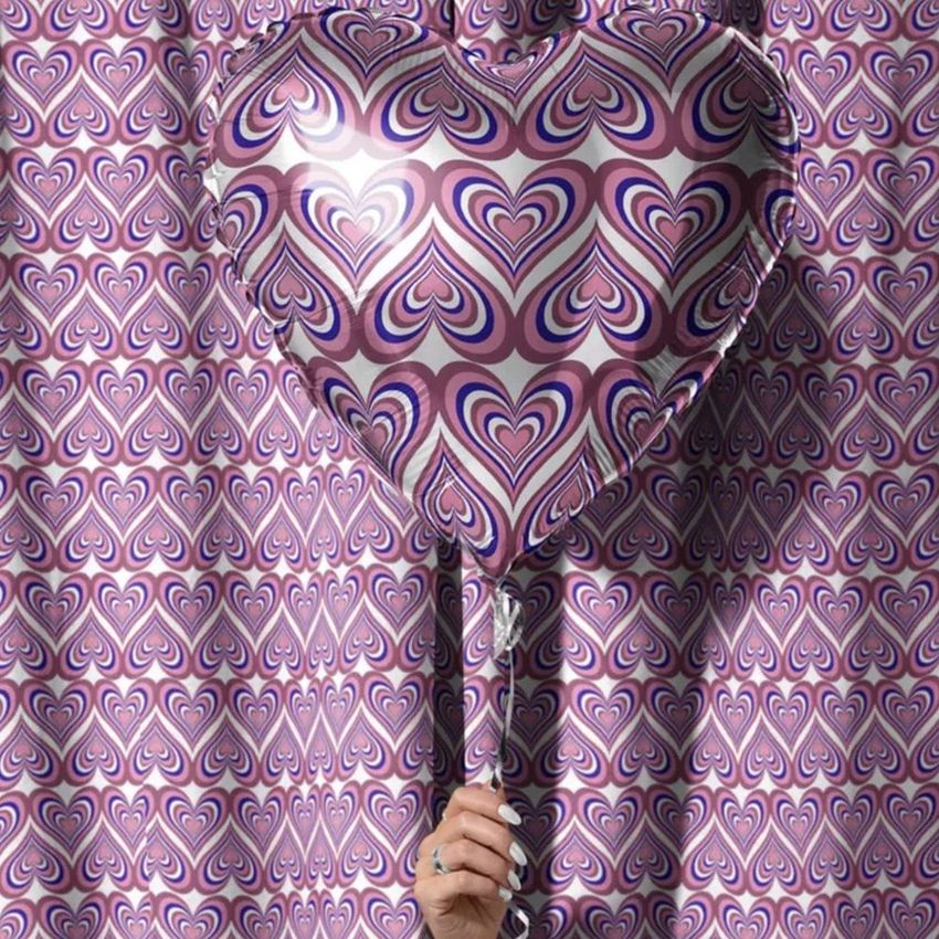 Hand holding a heart-shaped balloon with a retro purple and pink heart pattern, in front of matching velvet curtains.