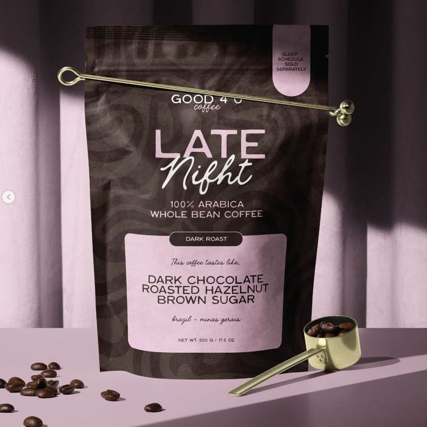 Coffee branding mockup featuring a dark coffee bag with pink and white text, gold scoop, scattered beans, and a mauve backdrop.