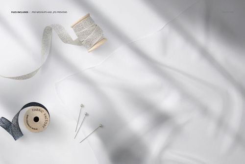 Close-up view of a square silk scarf mockup with two spools of ribbon and sewing pins on a light surface.
