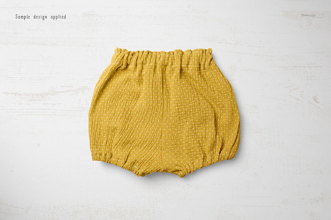 PSD mockup of baby shorts in mustard yellow with small white dots, laid out on a white wooden tabletop.