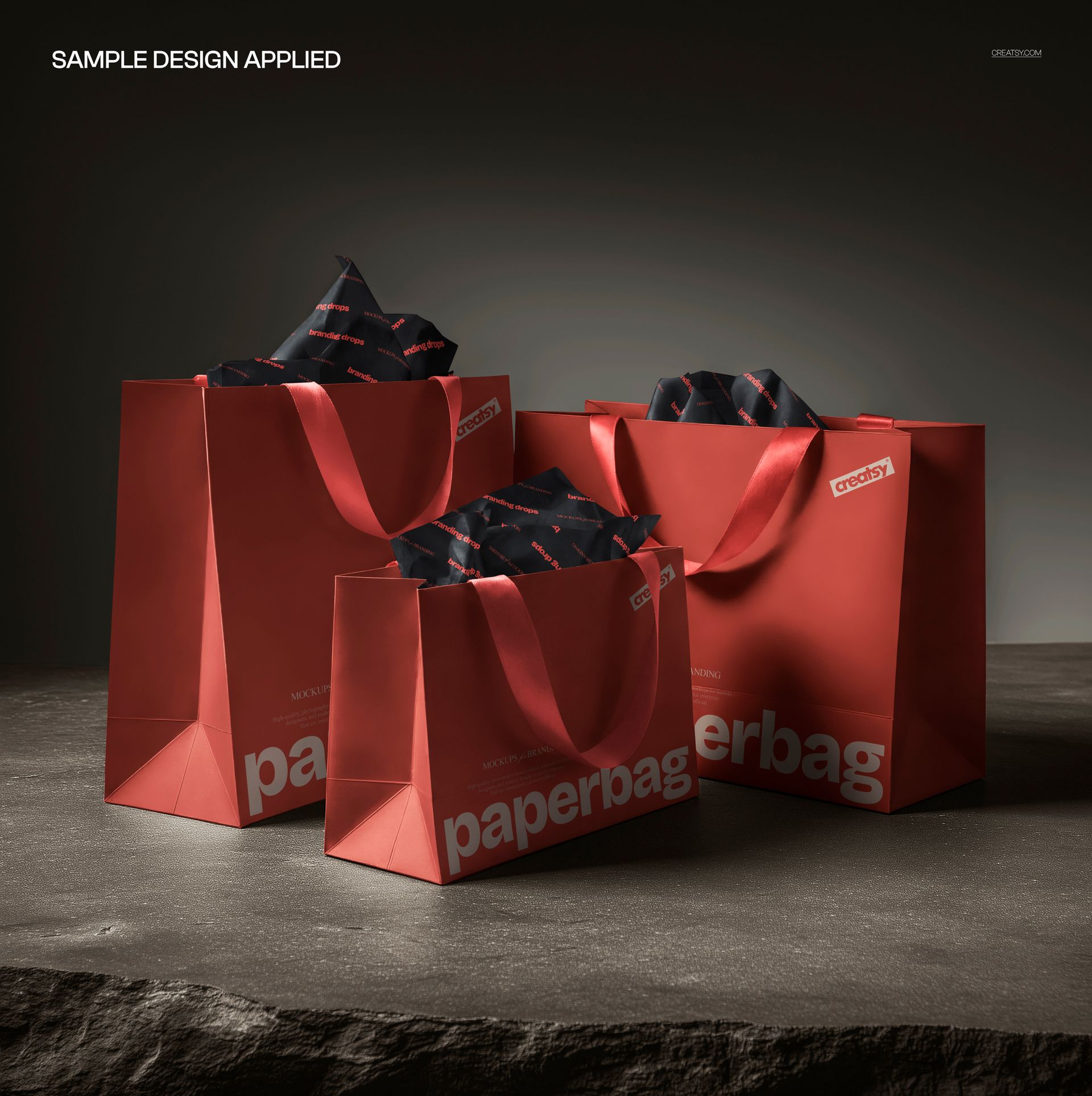 PSD mockup showing three red packaging bags with ribbon handles and patterned tissue paper on a stone background.