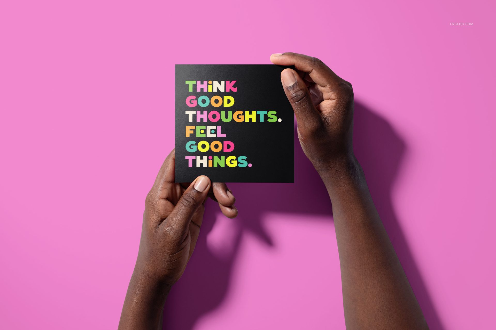 Folded square greeting card mockup featuring bold multicolored lettering, held by hands on a vibrant pink surface.