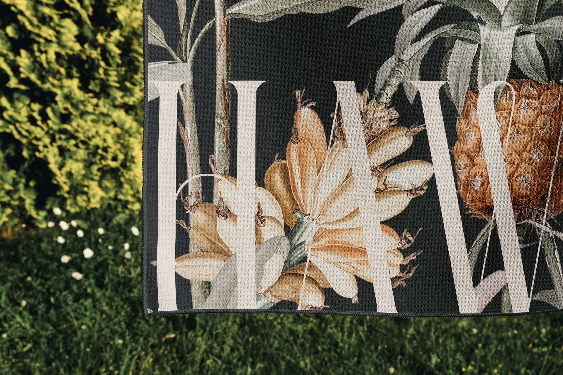 Close-up of a patterned waffle beach towel mockup featuring tropical flowers and fruit, displayed outside on grass.