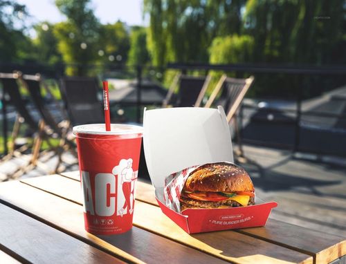 PSD mockup featuring a burger in packaging and a red paper cup with straw on a wooden table with chairs in background.