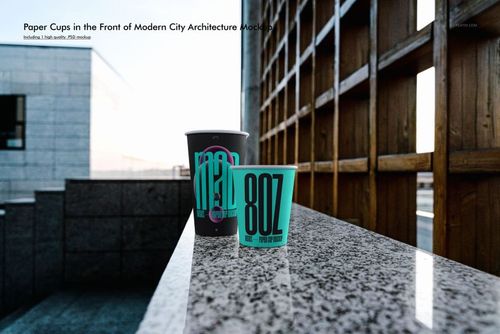 Paper cup PSD mockup showing two cups with vibrant text designs, positioned on a marble ledge near contemporary structures.