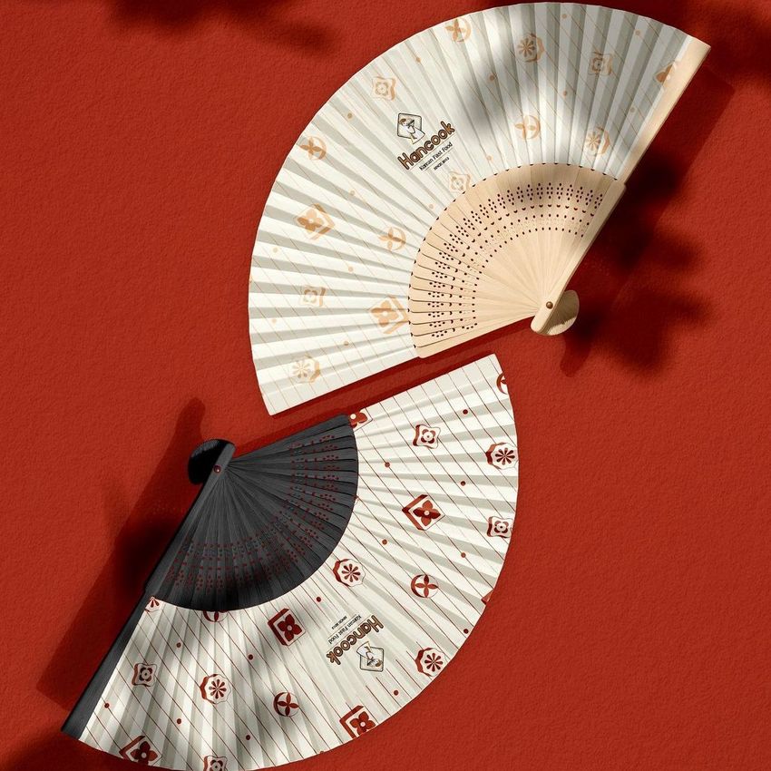 Fabric hand fan mockup showing two folding fans with patterned designs on a red textured surface, viewed from above.