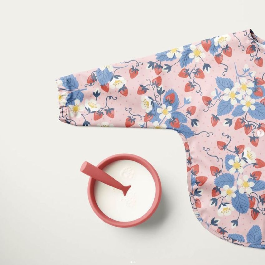 Baby sleeved bib mockup with a pink background and strawberry flower pattern, shown next to a red bowl of milk with a spoon.