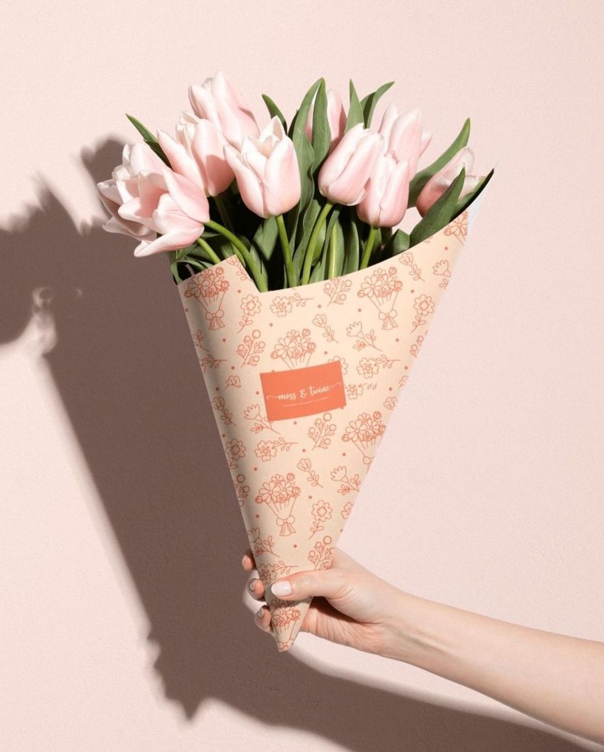 Hand holding a flower packaging cone mockup with pink tulips, featuring a floral pattern on beige paper against a light pink wall.