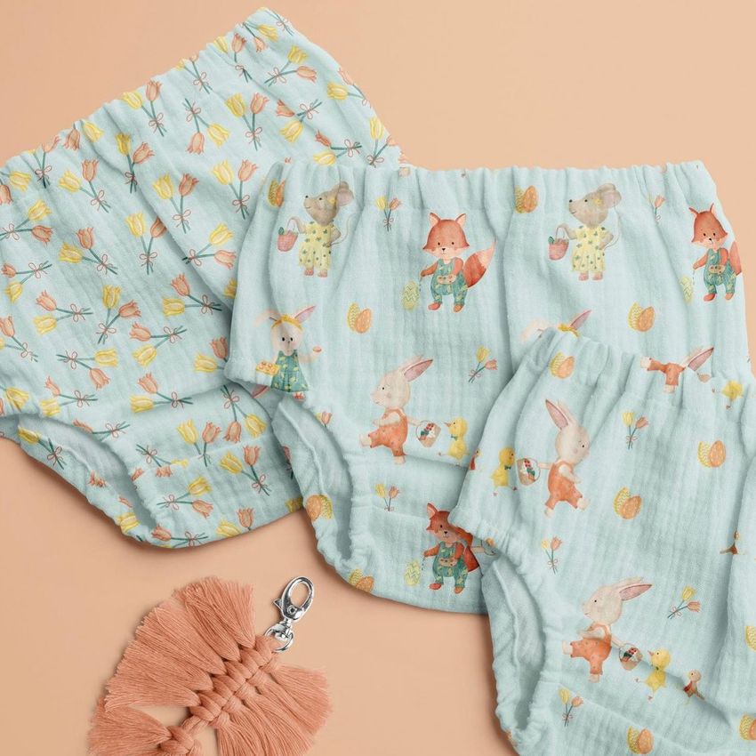 Baby muslin pants mockup featuring light blue fabric with woodland animal illustrations, shown on a peach background with a tassel keychain.