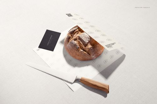 Mockup scene featuring a rustic bread loaf on branded food paper, knife with wooden handle, and minimalist background.
