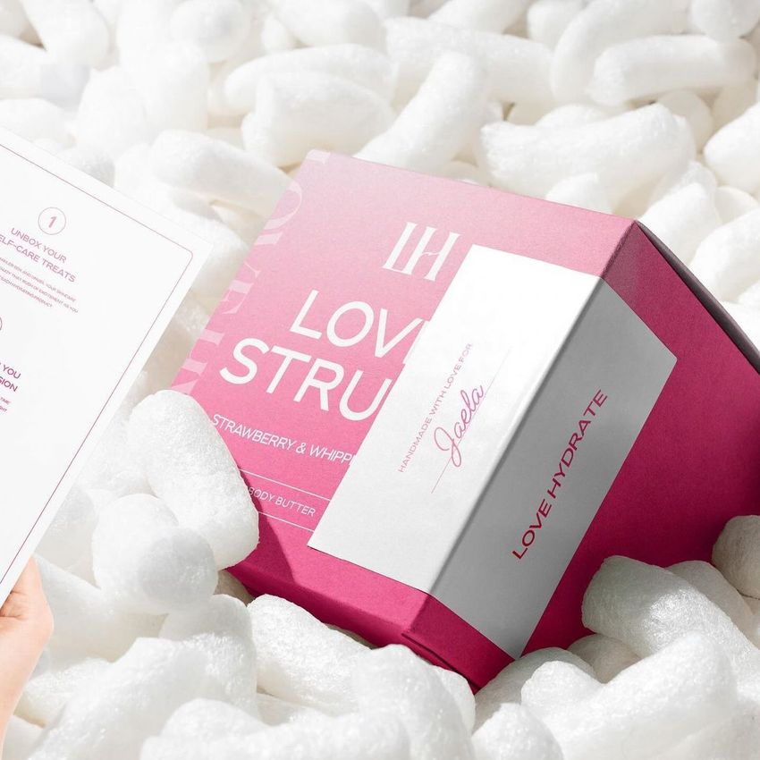Square box packaging mockup in pink and white placed among white packing peanuts, with a hand holding a paper insert nearby.