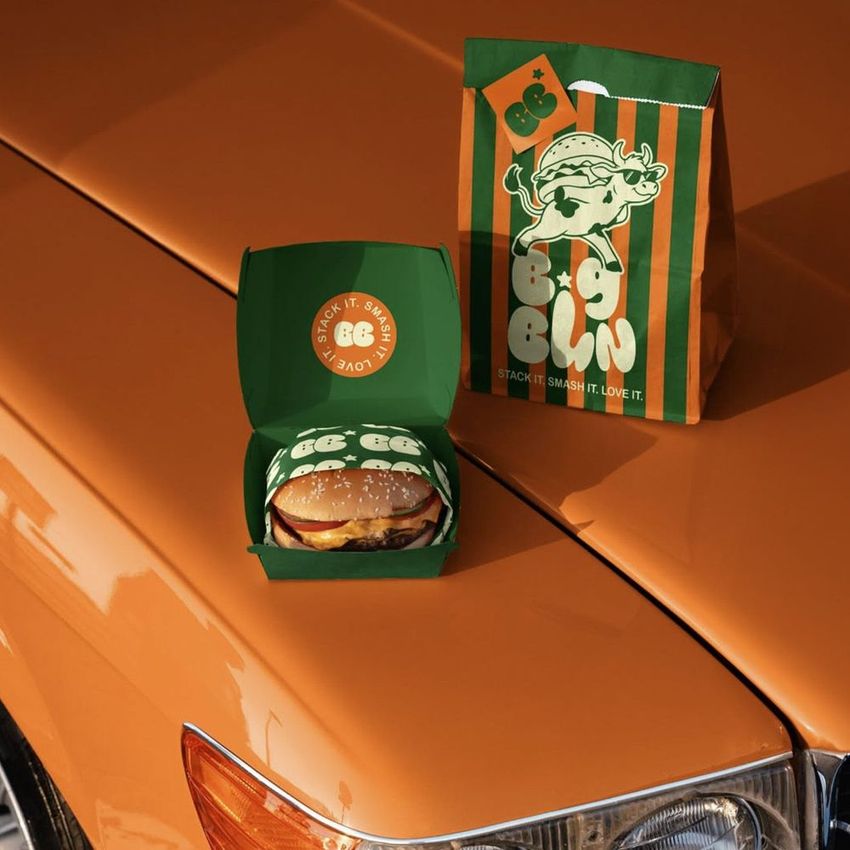 Burger packaging mockup displayed on the hood of a classic orange car, featuring a branded box and paper bag with illustrated graphics.