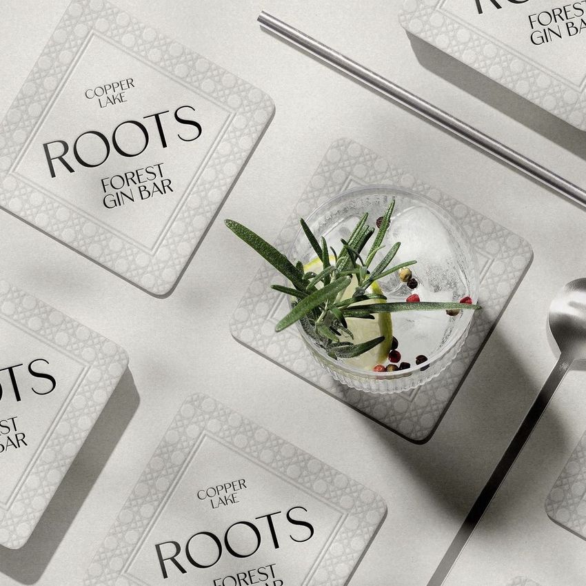 Square coaster mockup featuring several coasters with geometric border patterns, a gin cocktail, and metal bar tools on a light surface.