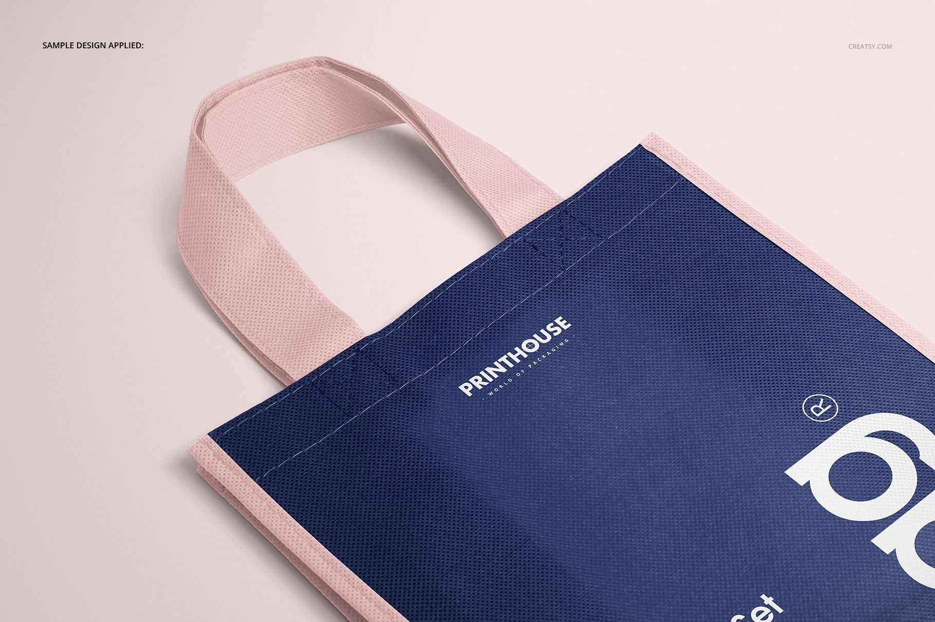 Close-up of a non-woven tote bag mockup with pastel pink handles and border, dark blue body, angled from above.