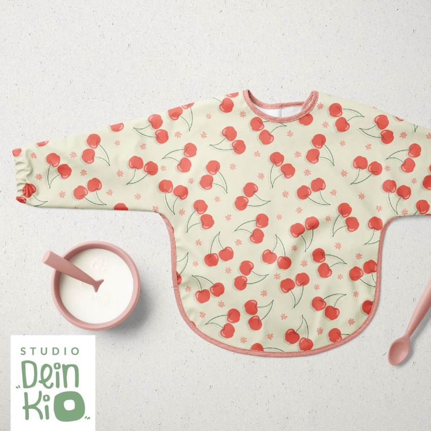 Baby sleeved bib mockup with a cherry and daisy pattern, shown flat on a light surface with a bowl, spoon, and text overlay.