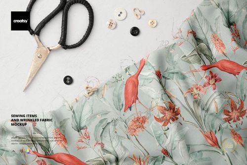 Top view of a PSD mockup showing wrinkled fabric with red birds and leaves, surrounded by sewing tools and buttons.