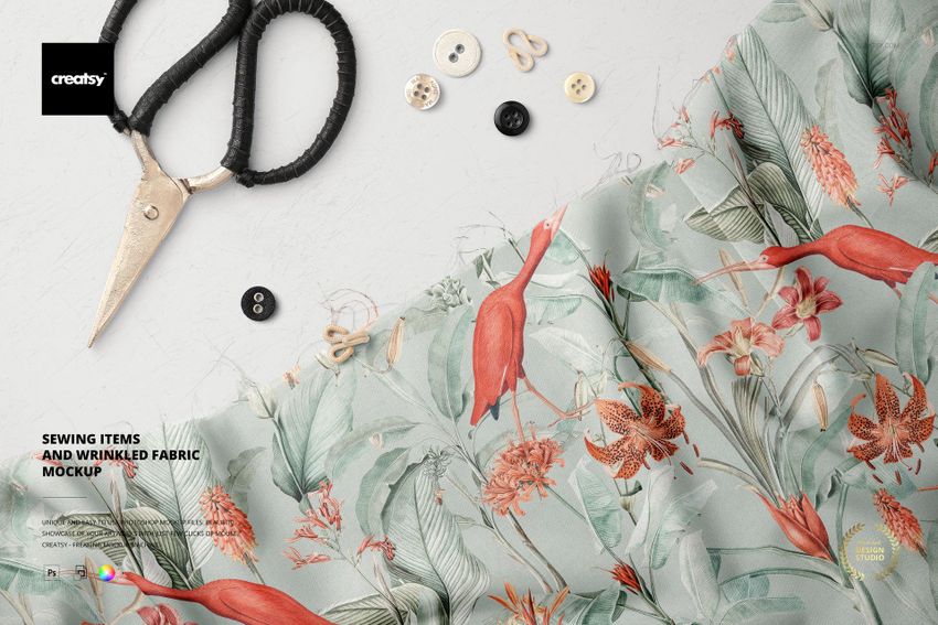 Top view of a PSD mockup showing wrinkled fabric with red birds and leaves, surrounded by sewing tools and buttons.