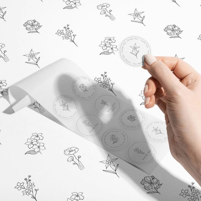 Round sticker mockup featuring a hand peeling a floral design sticker from a sheet on a white surface with flower sketches.