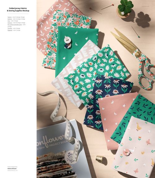 PSD mockup showing stacks of colorful jersey fabrics with floral, animal, and geometric designs, plus sewing accessories.