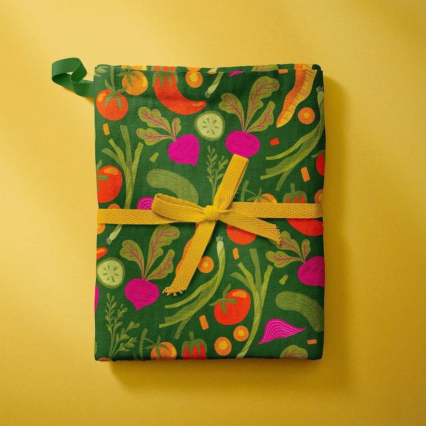 Kitchen towels mockup showing a folded green towel with colorful vegetable illustrations, tied with a yellow ribbon on a yellow background.