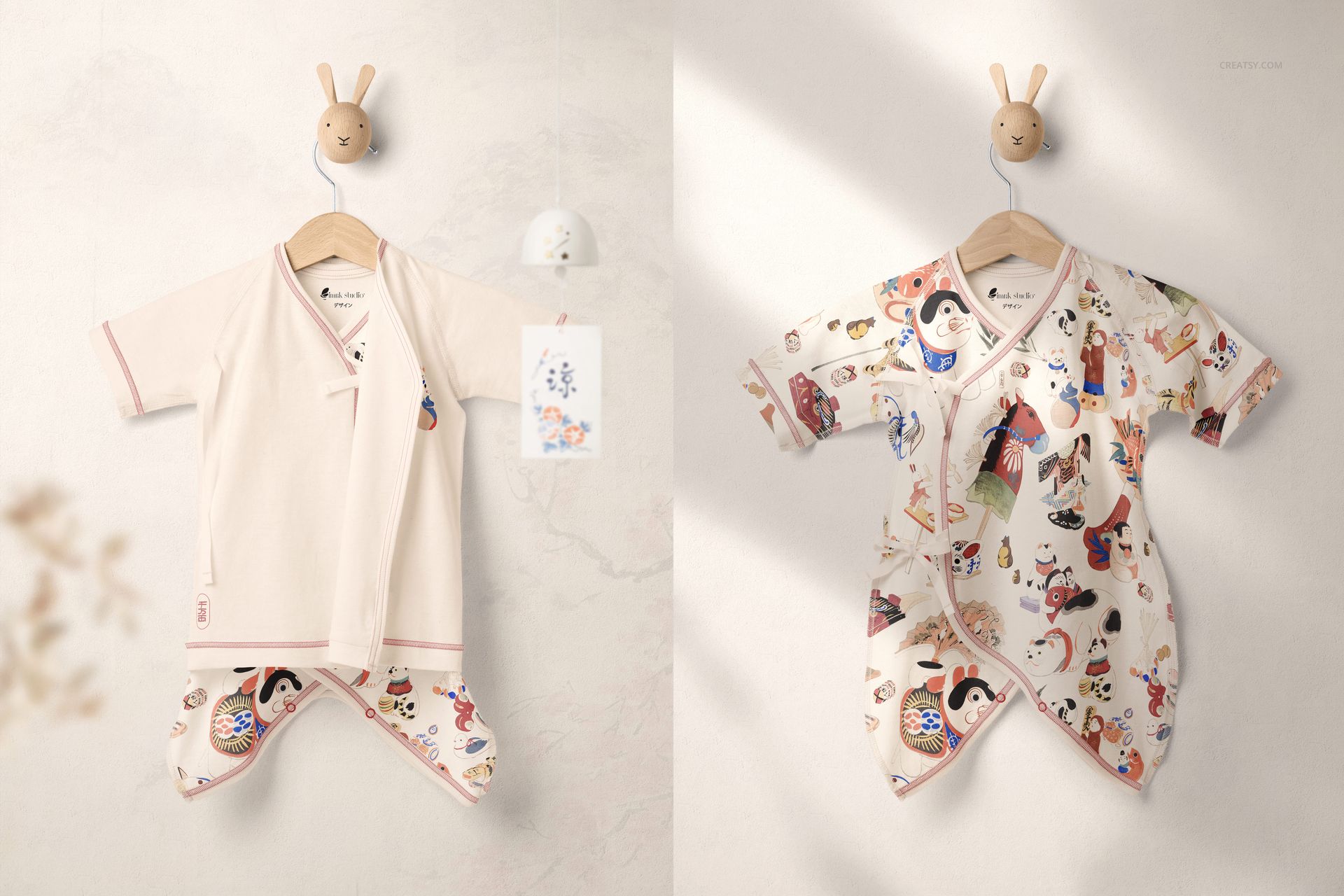 PSD mockup showing a pair of baby kimono bodysuits, one in neutral cream and one with vibrant traditional motifs, hanging on wall hooks.