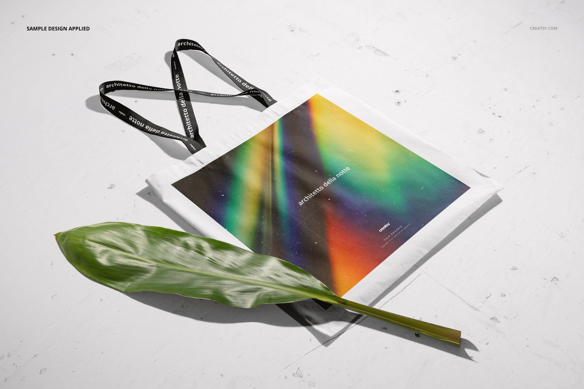 Tote bag mockup showing a colorful abstract print, black straps, and a large green leaf on a white background.