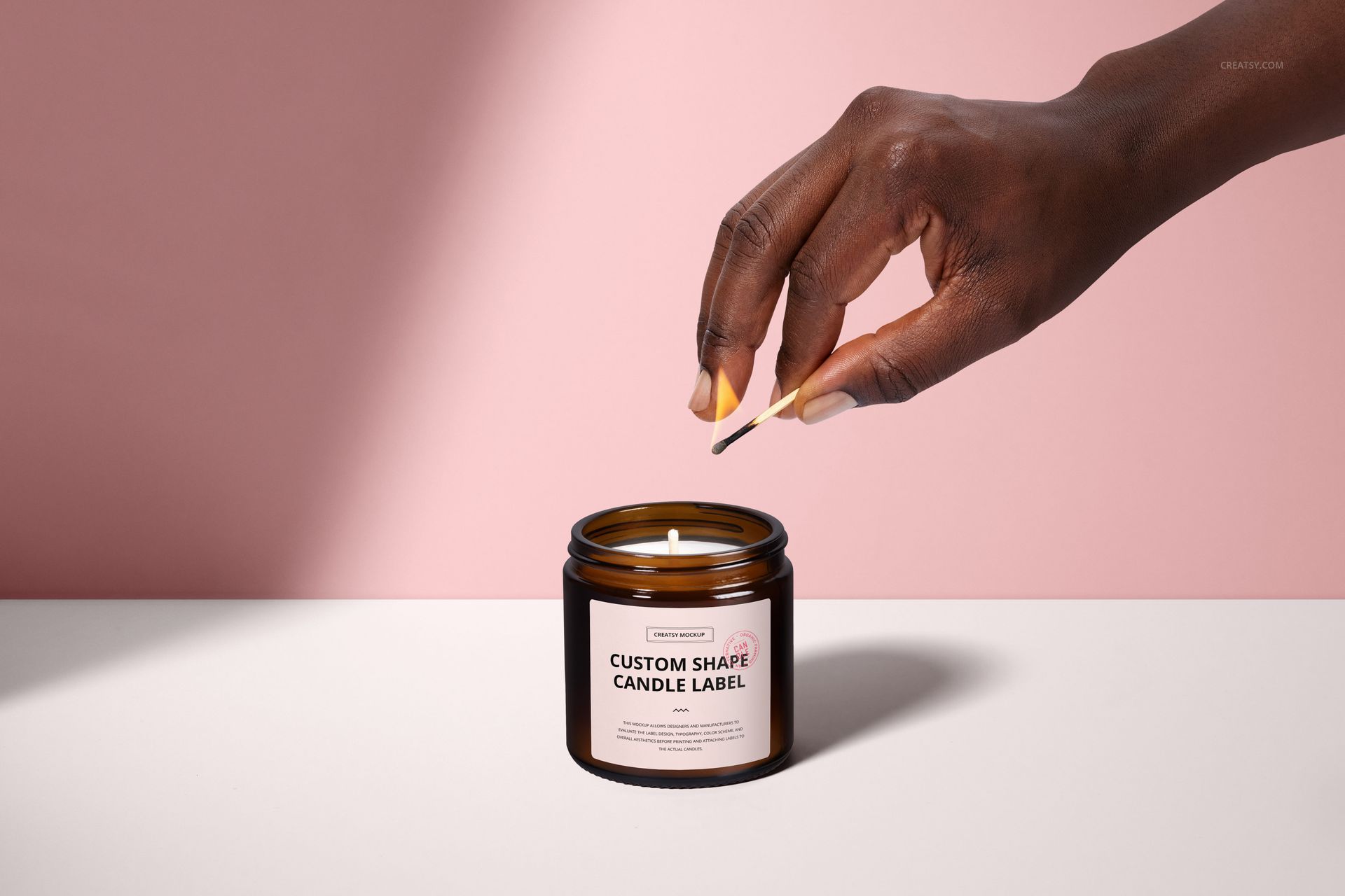 Front view of a brown amber jar candle mockup with cotton wick, hand holding match above, pastel pink wall.