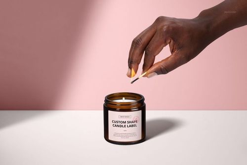 Front view of a brown amber jar candle mockup with cotton wick, hand holding match above, pastel pink wall.