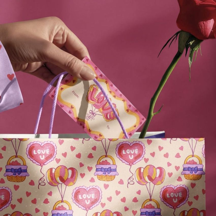 Gift packaging mockup showing a hand with a card and a rose being placed in a heart-themed bag with purple handles.