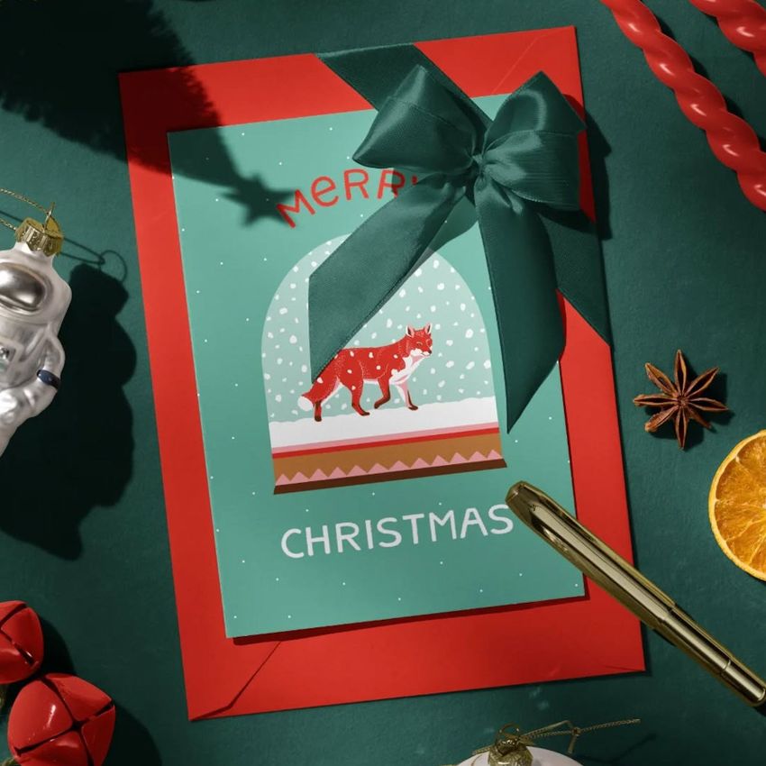 Christmas greeting card mockup featuring a red fox in a snow globe design, placed on a red envelope with a green ribbon bow.