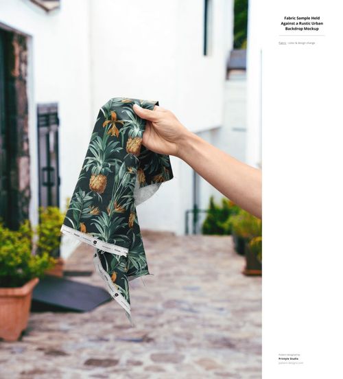 Fabric sample featuring a green and yellow tropical design, held up by hand in front of a rustic outdoor scene, PSD mockup.