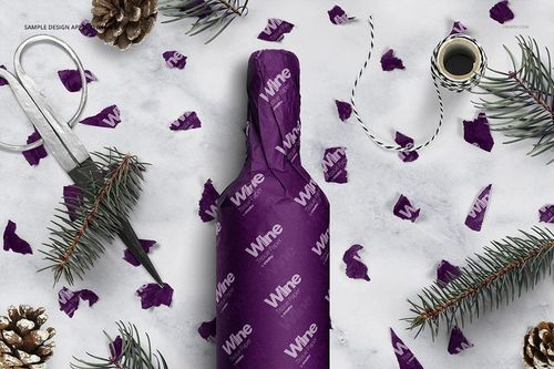 PSD mockup showing a wine bottle wrapped in purple tissue paper, with decorative pine branches and twine on a snowy background.