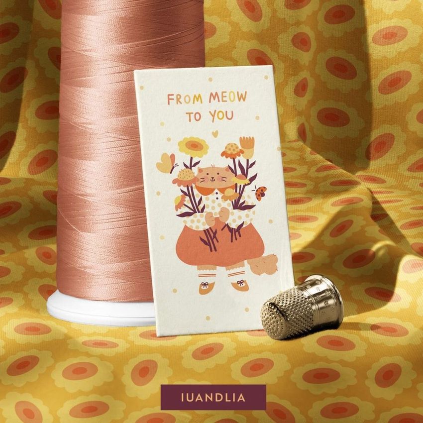 Thread spool and card mockup on patterned jersey fabric with a cute illustrated cat holding flowers, thimble nearby.