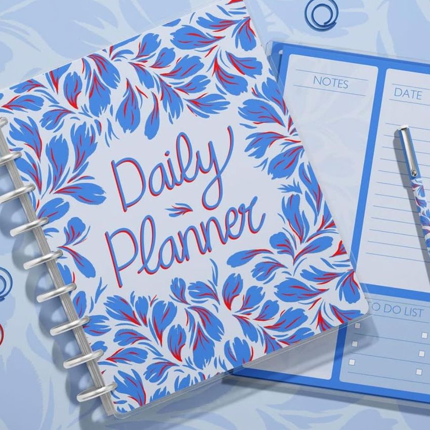 Top view of a spiral planner with a bold blue and red floral pattern, placed on a matching background next to a notepad and pen.