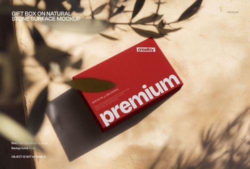 Front angled view of a red box mockup on a textured stone background, with leafy shadows cast across the scene.
