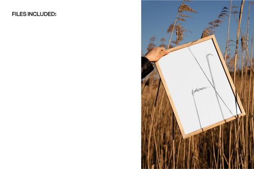 Wooden picture frame mockup held by hand among tall golden grass, with blue sky in the background.