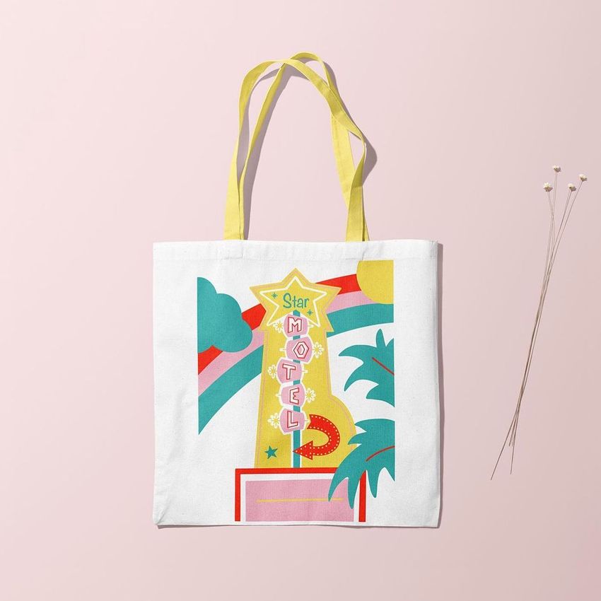 Tote bag mockup featuring a colorful retro motel sign illustration with palm trees, clouds, and geometric shapes on a light pink background.