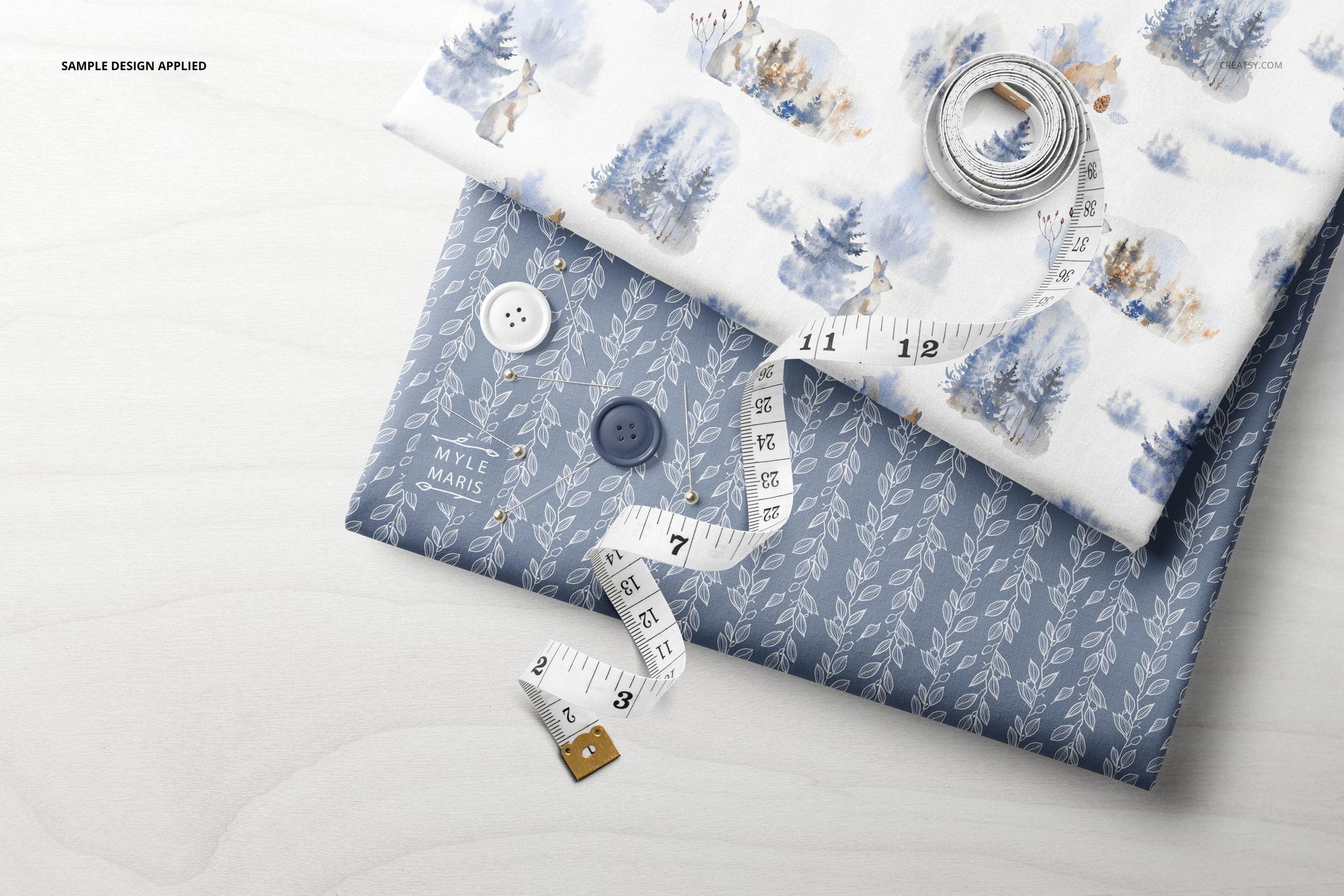 Textile mockup set featuring stacked folded fabrics with leaf and forest designs, accompanied by sewing accessories on a table.