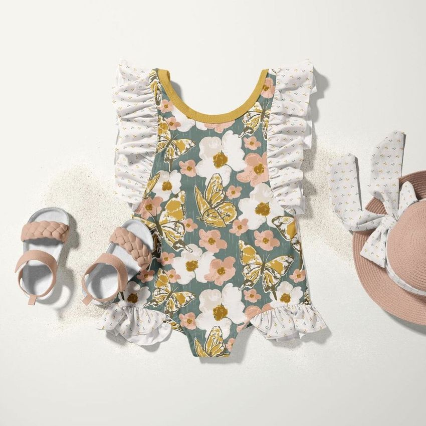 Kids swimsuit mockup with butterfly and floral pattern, ruffled sleeves, pink sandals, and matching sun hat on a light surface.