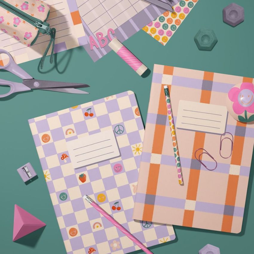 Stationery mockup featuring two classic A4 notebooks with colorful patterned covers, pencils, paper clips, and various desk accessories on a green surface.