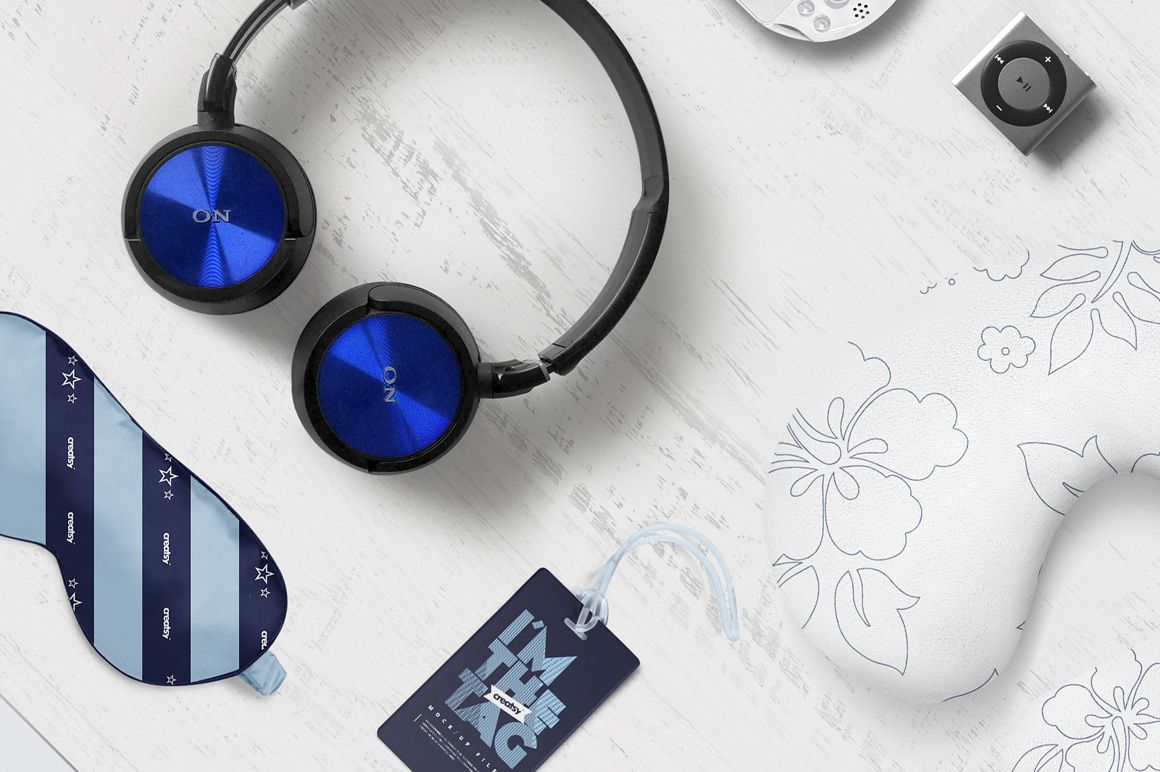 Travel identity mockup featuring blue headphones, striped sleep mask, floral neck pillow, and tag on white wood surface.
