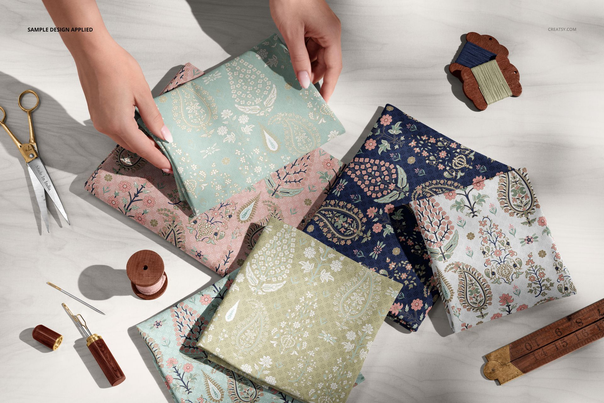 Hands arranging colorful jersey fabric pieces on a worktable with scissors, thread, and ruler, in a PSD mockup scene.