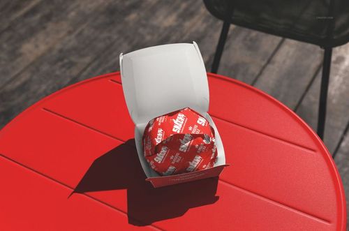 Fast food burger packaging mockup showing a white box with a red wrapped burger on a bright red table surface.