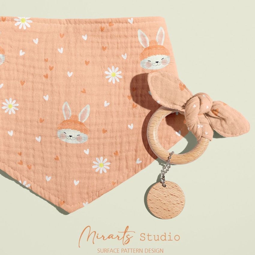 Baby muslin bandana mockup featuring a peach-colored fabric with cute rabbit faces and white flowers, paired with a wooden teether.