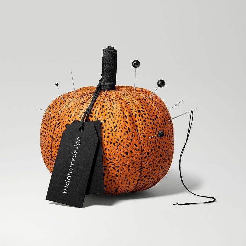 Fabric pumpkin mockup with an orange and black spotted pattern, black tag, and sewing pins on a plain gray background.