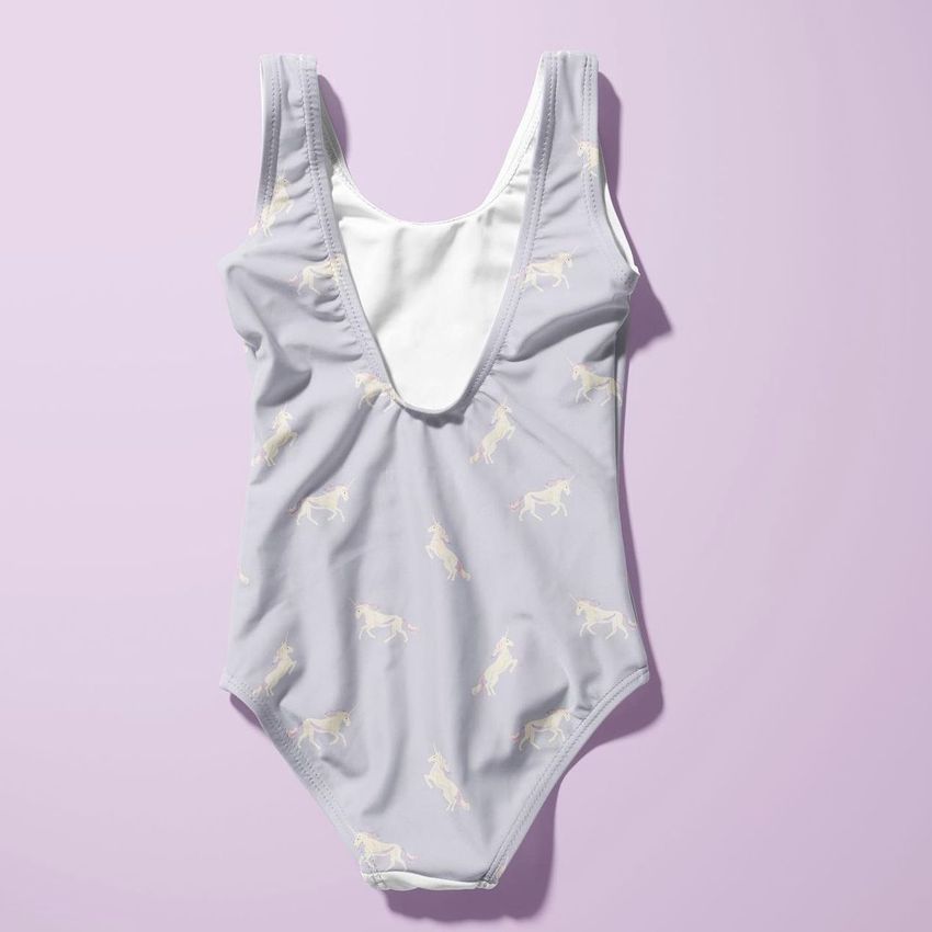 Baby swimsuit mockup featuring a sleeveless one-piece with a unicorn pattern on light fabric, displayed on a pastel purple background.