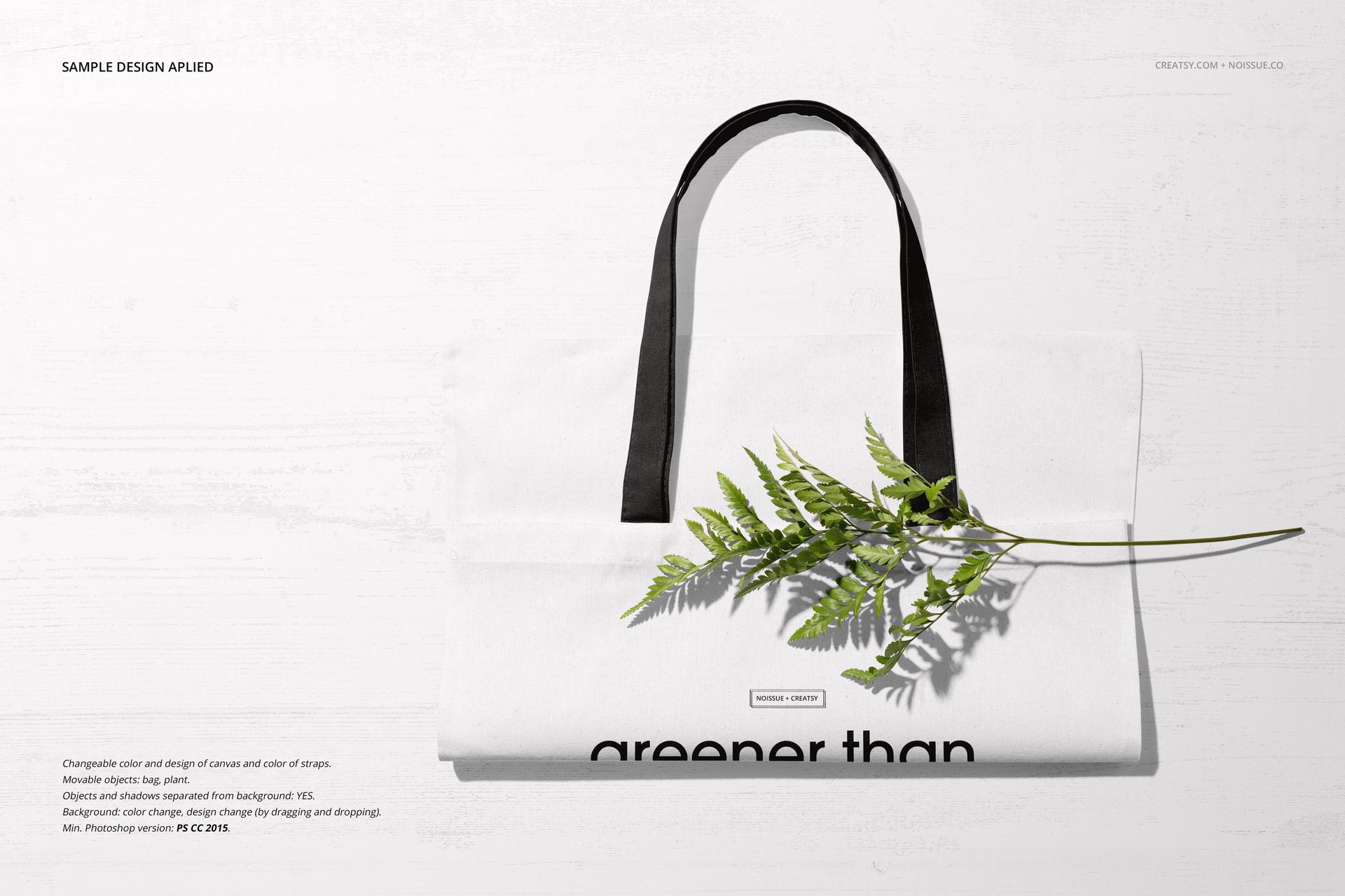 Front view of a white tote bag mockup with black handles, lying flat on a light wooden surface with a green fern branch.
