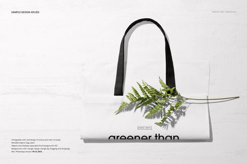 Front view of a white tote bag mockup with black handles, lying flat on a light wooden surface with a green fern branch.