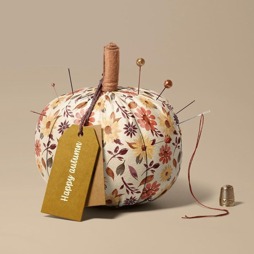 Fabric pumpkin mockup with a floral pattern, used as a pincushion, on a beige background with a gold tag and thimble.