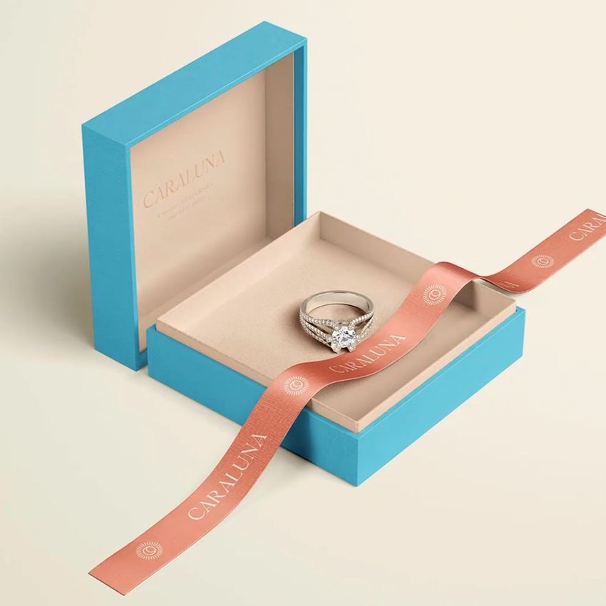 Jewelry box mockup featuring an open blue box with a ring inside, set on a light surface with a peach-colored ribbon and subtle text.